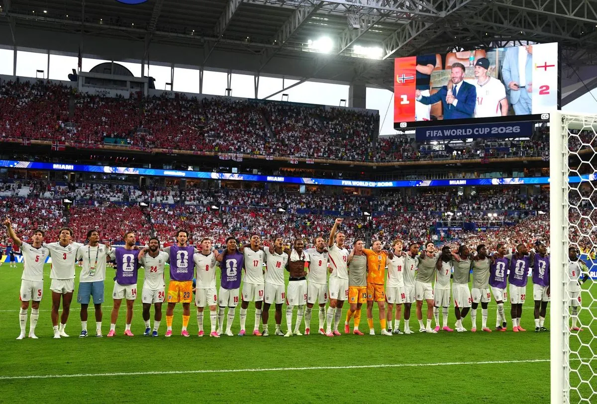 A group of athletes standing on a field in front of a large crowd, likely during a football match, with a large screen displaying a figure in the background.
