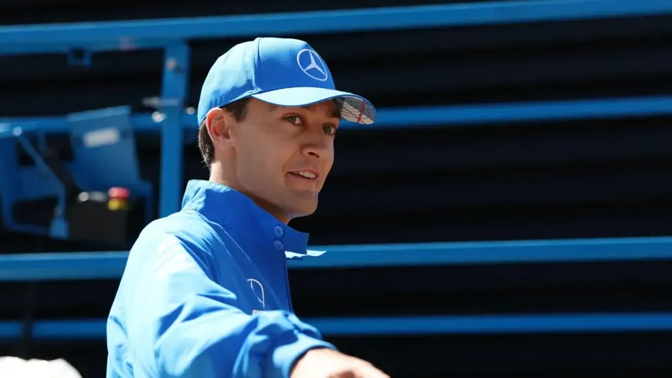 George Russell is wearing a bright blue baseball cap and a bright blue jacket. He is turning sideways and pointing.